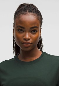 Camisa deportiva de color verde oscuro, de manga corta, con cuello redondo, tela texturada y un patrón sutil. Modelo con cabello trenzado y expresión neutral.