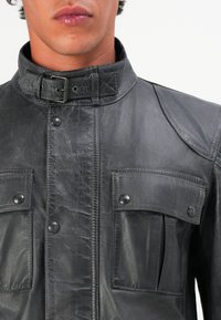 Chaqueta de cuero negra con cuello alto, collar abotonado, dos bolsillos en el pecho y acabado texturizado. Presenta botones a presión y costuras reforzadas.