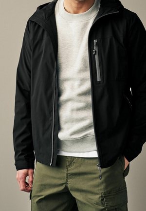 Mann trägt schwarze Reißverschlussjacke über hellgrauem Pullover, olivgrüne Cargohose, mit der rechten Hand in der Tasche und der linken Hand entspannt.