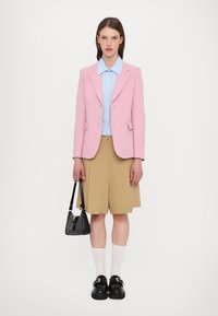 Blazer sartorial rosa, camicia azzurra chiara, pantaloni corti beige con plissettature, borsa nera e scarpe con plateau abbinate a calze. Sfondo semplice, design moderno.