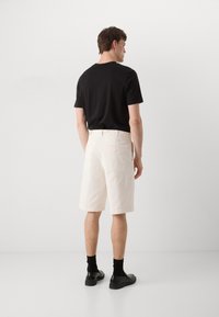 Weiße Baumwollshorts mit geradem Schnitt, ausgestattet mit Gesäßtaschen, getragen mit einem schwarzen Rundhals-T-Shirt, schwarzen Socken und dunklen Schuhen.