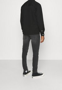 Zwart overhemd met knopen en een achterpandnaad, gecombineerd met nauwsluitende grijze jeans en zwarte hoge sneakers, die een casual stijl uitstralen.