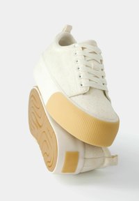 Beżowo-kremowe sneakersy na platformie z teksturowaną tkaniną, grubymi beżowymi żebrowanymi podeszwami, białymi sznurowadłami i pętlami do zakładania na białym tle.