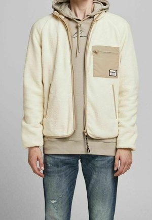 Mann, der eine cremefarbene Fleecejacke mit einer hellbraunen Reißverschlusstasche auf der Brust über einem beigen Hoodie und blauen, abgenutzten Jeans trägt.