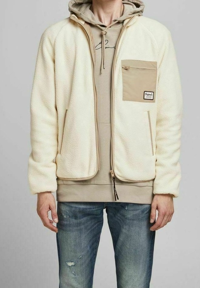 Homme portant une veste polaire de couleur crème avec une poche poitrine zippée beige par-dessus un sweat à capuche beige et un jean bleu déchiré.