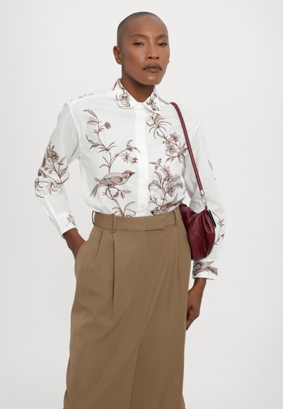 Camasa albă cu broderie florală maro, asortată cu pantaloni bej cu talie înaltă și o geantă mică de culoare vișinie. Texturi netede.