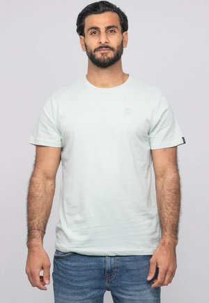 Homme aux cheveux foncés et à la barbe portant un t-shirt bleu clair et un jean bleu, debout devant un fond gris uni.