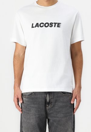 Biała koszulka z krótkim rękawem z logo "LACOSTE" czarnymi literami na piersi, noszona z wyblakłymi czarnymi dżinsami.