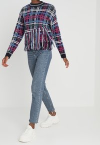 Gestrickter Pullover mit einem mehrfarbigen Karomuster und einem fransigen Saum. Kombiniert mit eng anliegenden blauen Jeans und weißen Turnschuhen.