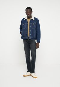 Jeans jakna s bež flis ovratnikom, svetlo modra, z gumbi na sprednji strani, v kombinaciji s temnimi kavbojkami in belimi supergami. Model stoji ob beli podlagi.