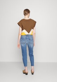 Hnědý svetr s krátkým rukávem a výstřihem ve tvaru V, přehozený přes žlutou košili. V kombinaci se světle modrými džínami a béžovými botami na blokov ém podpatku.