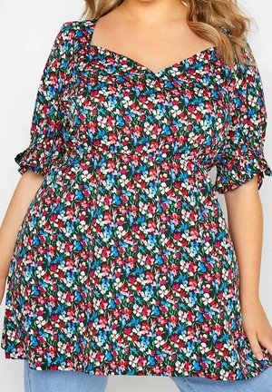 Femme grande taille portant une robe noire à fleurs rouges, blanches, bleues et roses avec des manches bouffantes et des poignets à volants.