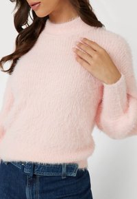Helles rosa, flauschiges Strickpullover mit hohem Kragen, geripptem Muster und leicht übergroßen Ärmeln, kombiniert mit dunkler Jeans.