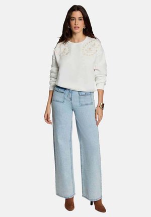 Femme portant un sweat-shirt blanc avec des détails floraux légers, un jean large bleu clair avec des poches avant, et des bottines marron à talons.