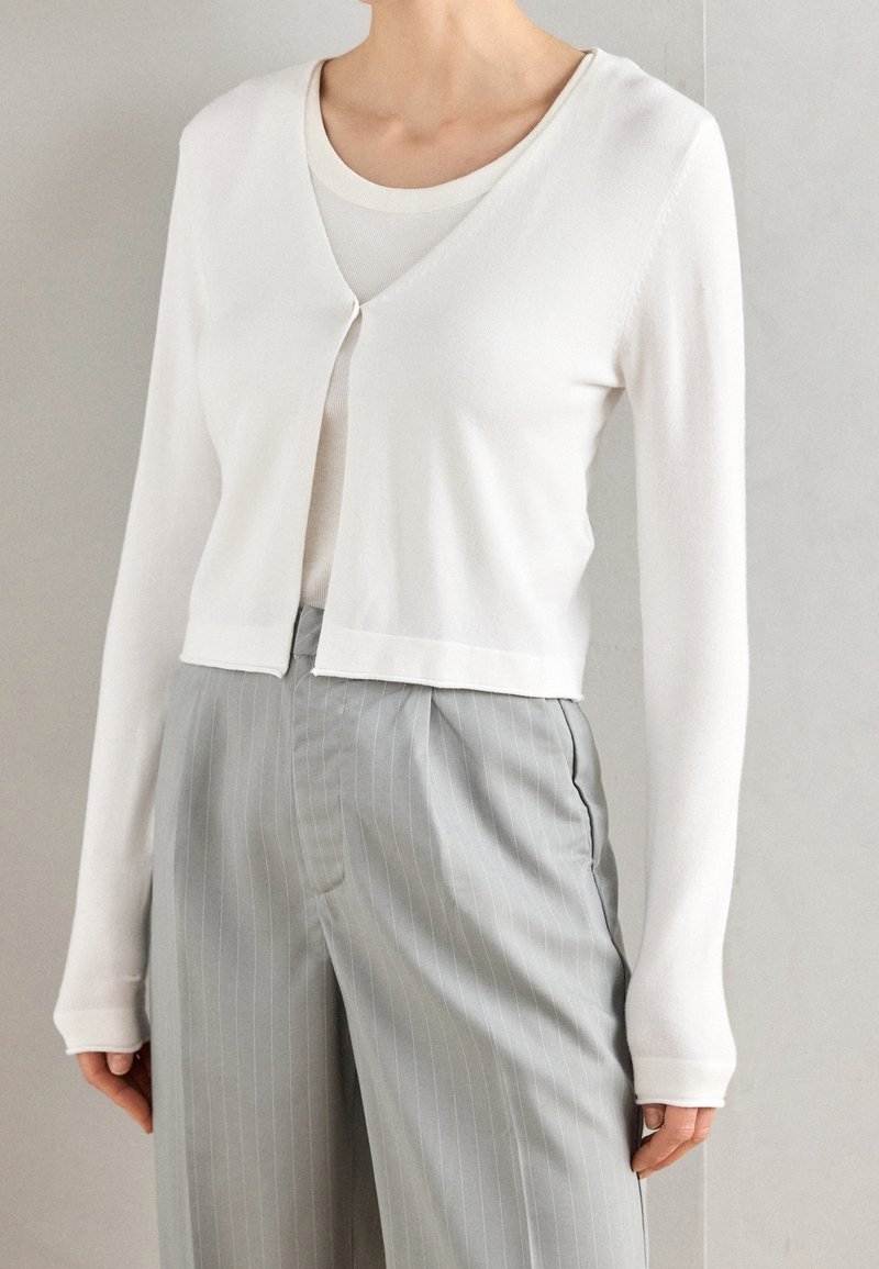 Femme portant un cardigan blanc court sur un haut blanc à encolure dégagée, associé à un pantalon gris à fines rayures taille haute sur un fond uni.