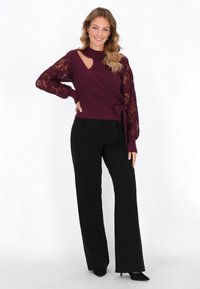 Burgundin värinen ribattu neule, jossa on pitsihihat, leikkauksellinen yksityiskohta ja sidottu vyötärö, yhdistettynä mustiin leveälahkeisiin housuihin ja mustiin korkokenkiin.