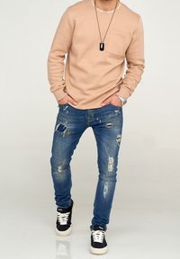 Beige långärmad skjorta med ficka och ribbad halsringning, i kombination med slitna blå jeans som har revor och färgstänk. Svarta sneakers.