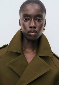 Close-up of serious woman with short hair wearing olive green coat and large gold hoop earrings against plain background.