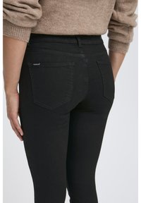 Jean en denim noir slim fit, doté de deux poches arrière et de détails de couture subtils. Le tissu a une texture lisse.