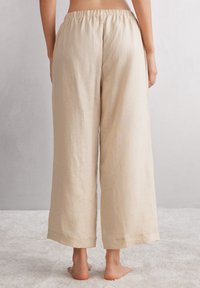 Beige Leinen-Hosen mit weitem Bein und elastischem Bund, die einen lockeren Schnitt und gesäumte Bündchen aufweisen, vor einem sanften, neutralen Hintergrund.