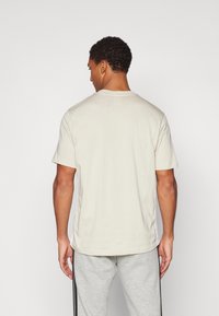Beige T-Shirt mit kurzen Ärmeln, Rundhalsausschnitt und lockerer Passform, aus leichtem Baumwollstoff, von hinten vor einem weißen Hintergrund gezeigt.