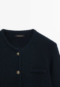 Cardigan en maille texturée bleu marine foncé avec encolure ronde, deux boutons métalliques et une petite poche poitrine côté droit.