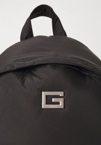 Juodas kuprinis krepšys su blizgiu sidabriniu "G" logotipu. Medžiaga atrodo lygi su šiek tiek blizgesio. Yra užtrauktuko viršus ir sukilęs rankena.