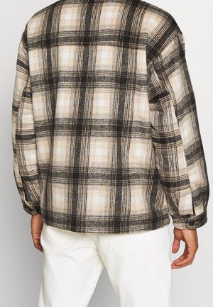 Camisa de franela a cuadros con un patrón en negro, crema y beige. Presenta un corte relajado y puños abotonados. La vista trasera muestra un detalle de yugo.