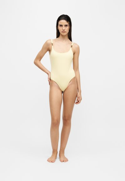 Femme debout pieds nus portant un maillot de bain une pièce jaune pâle à fines bretelles sur un fond clair uni.