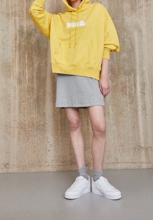 Personne portant un sweat à capuche jaune Nike, une jupe grise, des chaussettes grises et des baskets blanches Nike, debout contre un mur en béton texturé.