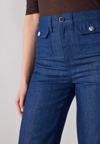 Ψηλόμεσα σκούρα μπλε τζιν παντελόνια με μπροστινές τσέπες, μεταλλικές λεπτομέρειες και λεία υφή, διαθέτοντας κλασικό σχέδιο ίσιας γραμμής.