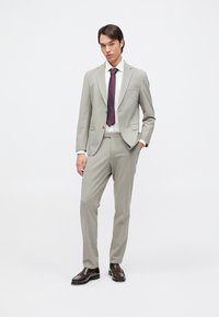 Young man standing in light gray suit with white shirt, dark red tie, and dark brown dress shoes against white background.