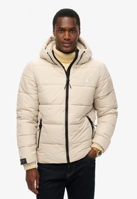 Beige pufferjakke med hætte, sorte lynlåse og forlommer. Har et quiltet design og ribbet krave. Accompanyeres af en cremet sweater.