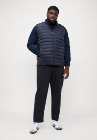 Veste rembourrée navy avec un col montant, fermeture éclair et poches latérales. Portée sur une chemise navy à manches longues et associée à un pantalon noir et des baskets.