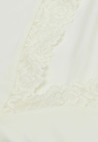 Krämfärgad tyg med spetskant, som har en mjuk textur och blommönster längs kanten. Mjukt, lätt material med en delikat finish.