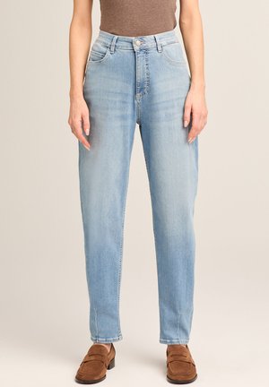 Woman wearing light blue high-waisted straight-leg jeans, brown top, and brown suede loafers, standing against a plain background.