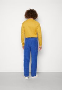 Sweat-shirt jaune à col rond, associé à un pantalon cargo en velours côtelé bleu avec des poches. Des baskets blanches complètent la tenue.