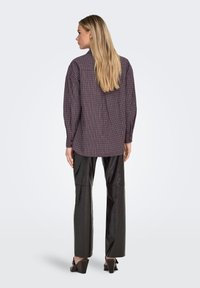 Blouse à carreaux violet foncé et crème, coupe ample, manches longues, associée à un pantalon en cuir noir brillant et des talons compensés.