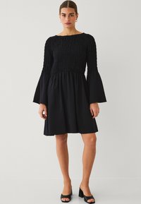 Zwarte jurk met een gesmokt bovenlijfje, lange klokmouwen en een flared rok. De stof lijkt textuur te hebben, wat visuele interesse creëert.