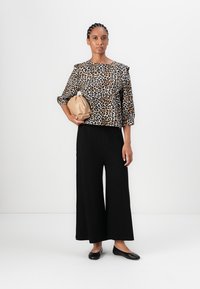 Top con stampa leopardata e maniche a tre quarti, abbinato a pantaloni neri a gamba larga. Tiene una borsa a mano beige e indossa delle scarpe flat nere.