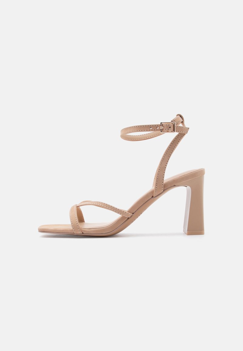 Beżowy damski sandał na szerokim obcasie, z dwoma cienkimi paskami na palcach oraz paskiem wokół kostki z klamrą, na białym tle.