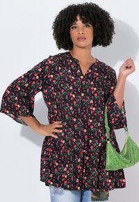 Tunique noire à fleurs avec des manches évasées, présentant un motif lumineux de fleurs roses et oranges. Associée à un sac à main texturé vert.