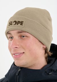 Beige gerippte Mütze mit einem braunen "SLOPE" Logo-Patch. Der Stoff sieht weich aus, und die Mütze sitzt eng am Kopf.