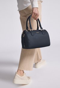 Donkerblauwe leren handtas met een gestructureerde afwerking, gouden hardware, dubbele handvatten en een bagagetag. Draagt bij beige broeken en witte sneakers.
