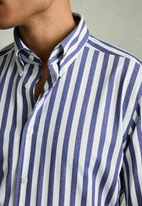 Camisa de botones a rayas azul y blanca con cuello clásico, fabricada en una mezcla de algodón y con detalles de botones visibles en la tapeta.