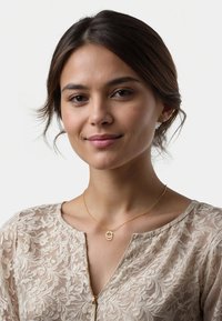 Guld halskæde med en cirkulær vedhæng båret over en gennemskinnelig bluse i blomstermønster i lys beige. Glat tekstur og subtil glans på materialerne.