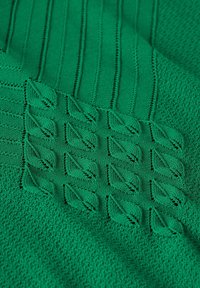 Grön stickad tyg med vertikala ribbor och texturerade bladmönster. Materialet är mjukt med en något upphöjd design som skapar visuell intresse.