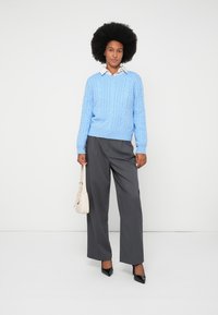 Nainen, jolla on kihara hius, pukeutuneena vaaleansiniseen palmikkoneuleeseen, harmaisiin leveälahkeisiin housuihin, mustiin korkokenkiin ja hän pitää valkoista käsilaukkua valkoista taustaa vasten.