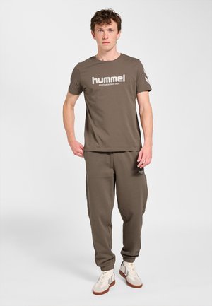 Mężczyzna w brązowej koszulce Hummel i dopasowanych dresach oraz białych trampkach, stojący na jednolitym białym tle.