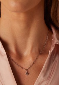 Zilveren ketting met uitgerekte schakels, met een metalen "N" hanger die centraal hangt. Draag het over een lichtroze blouse.